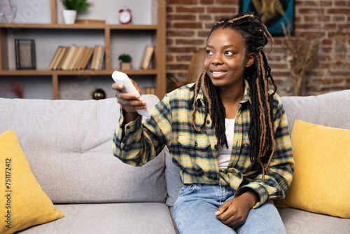 Content African American woman relaxing on couch, utilizing remote control for air con at a comfortable and stylish living room. Experiencing home comforts and smart living.
