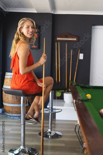 Young Caucasian woman in an orange dress enjoys a game of pool, holding a cue stick with copy space