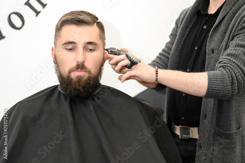 A barber cuts a man with a beard in a barber shop. Short haircut of the client with clipper.