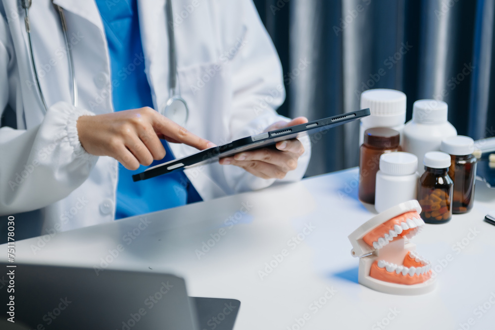 Dentist sitting at table with jaw samples tooth model and working with tablet and laptop in dental modern office Dentist sitting at table with jaw samples tooth model and working with tablet and laptop in dental modern office