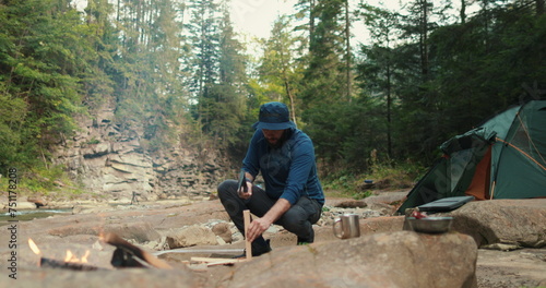 a male tourist chops firewood with a small ax near a campfire in the forest. the traveler prepares wood for the fire. man in the wild. outdoor hiker, wilderness survival, campfire