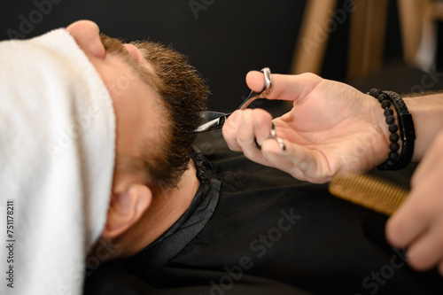 A barber stylist trims the beard of a Caucasian man, whose face is covered with towel, with scissors