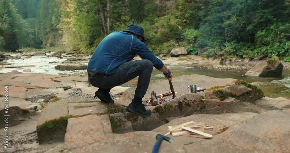 a male tourist chops firewood with a small ax near a campfire in the forest. the traveler prepares wood for the fire. man in the wild. outdoor hiker, wilderness survival, campfire a male tourist chops firewood with a small ax near a campfire in the forest. the traveler prepares wood for the fire. man in the wild. outdoor hiker, wilderness survival, campfire