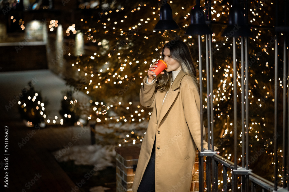 beautiful young girl in a beige coat with a paper glass of coffee against the backdrop of many bokeh lights beautiful young girl in a beige coat with a paper glass of coffee against the backdrop of many bokeh lights