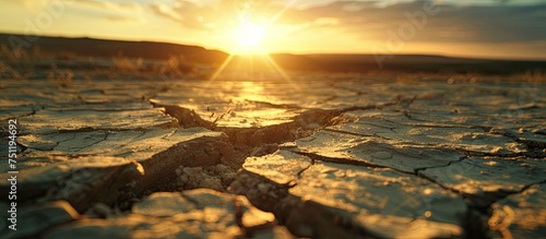 The sun is low on the horizon, casting a warm golden glow over a cracked road. The fissures in the dry land create a stark contrast against the setting sun.