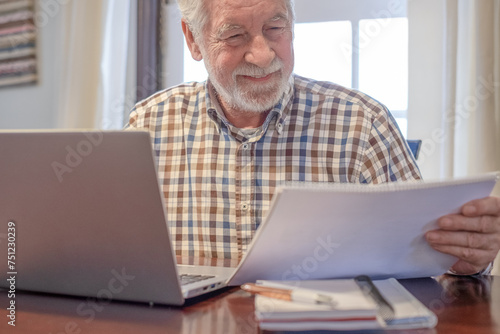 Smiling bearded man sitting at table with laptop and books working or studying. Attractive senior man enjoying teaching activity and learning using new technology photo