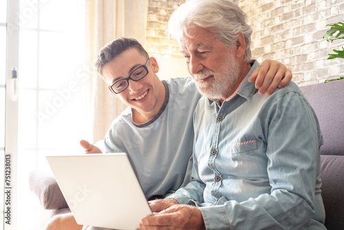 Smiling experienced young boy with laptop teaching and showing new computer technology to his old grandfather for surfing internet, shopping online, managing bank. Old man learns to use the computer photo