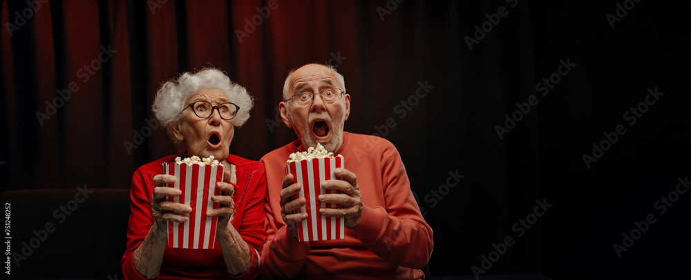 Bright facial expression, human emotions concept. Funny portrait of two senior old couple scared shocked or impressed group with popcorn in hands. Enjoy watching horror movie or thriller in the cinema