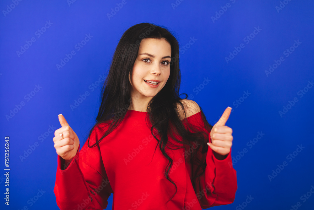 Optimistic brunette hair woman makes thumbs up gives approval shows excellent gesture says nice job gives positive feedback shares good opinion, wow, wear red top isolated over blue background. Optimistic brunette hair woman makes thumbs up gives approval shows excellent gesture says nice job gives positive feedback shares good opinion, wow, wear red top isolated over blue background.