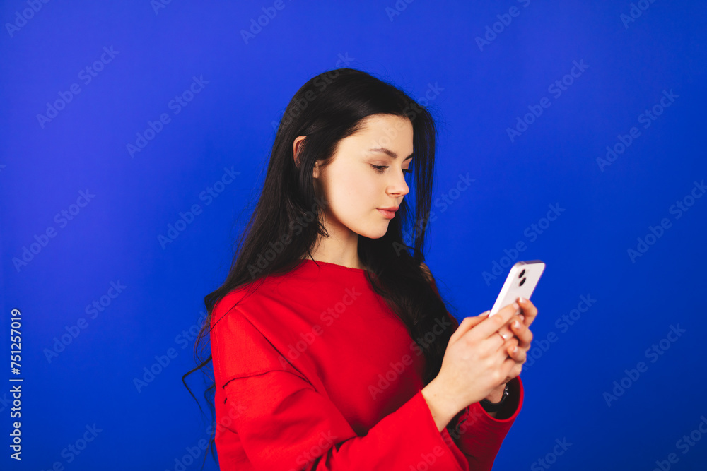 Indoor shot of serious woman uses modern gadgets uses mobile phone, girl wear red long sleeves top sends sms, watch social media message isolated on blue background. Indoor shot of serious woman uses modern gadgets uses mobile phone, girl wear red long sleeves top sends sms, watch social media message isolated on blue background.