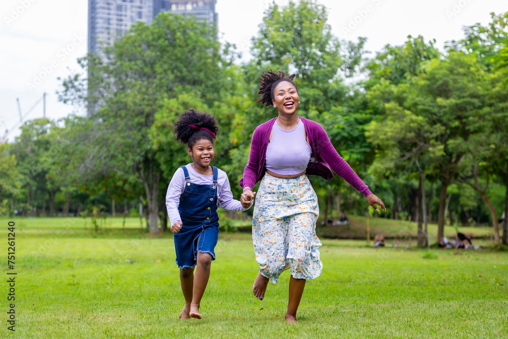 African American mother running barefoot on the grass lawn with her young daughter while having a summer picnic in the public park for wellbeing and happiness concept