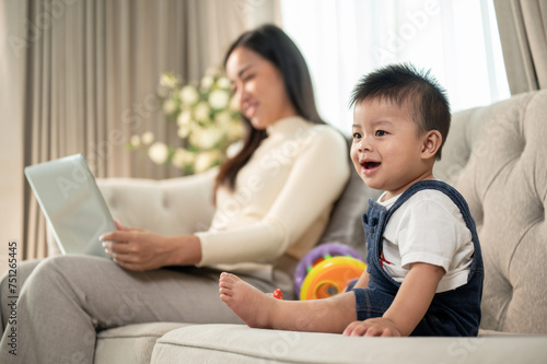 A cute Asian baby boy is playing with toys on a sofa while his mom is working on her laptop.