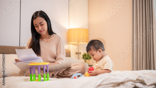 A cute Asian baby boy is playing with toys in bed while his mom is focusing reading documents.