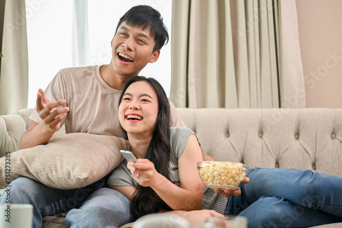 A happy young Asian couple is relaxing on a sofa, enjoying watching television together at home.