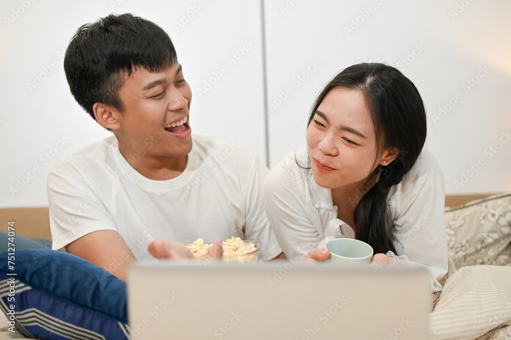 Happy Asian couple is having a fun time in bed, enjoying popcorn while watching a movie on a laptop. Happy Asian couple is having a fun time in bed, enjoying popcorn while watching a movie on a laptop.