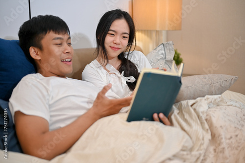 A happy young Asian couple in pajamas enjoys reading a book together before going to bed.