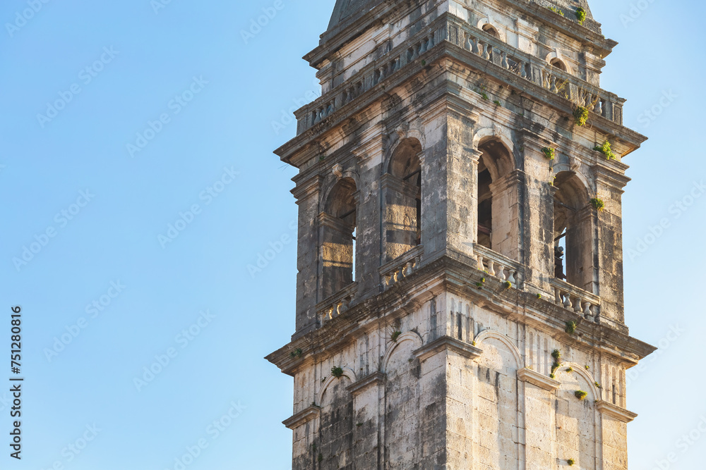 Weathered bell tower rises against clear blue sky in Perast, Montenegro; aged stone, arched openings, vegetation sprouting from crevices, baroque architecture. Close up, copy space