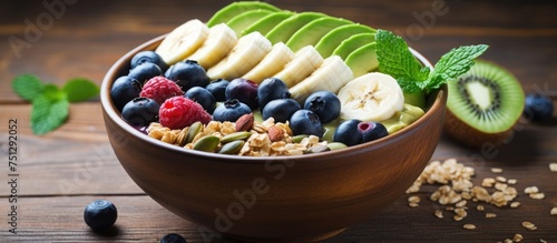 A bowl placed on a wooden table is filled with an assortment of fruits and nuts, including blueberries, mulberries, bananas, avocado, spinach, granola, coconut, and kiwi.