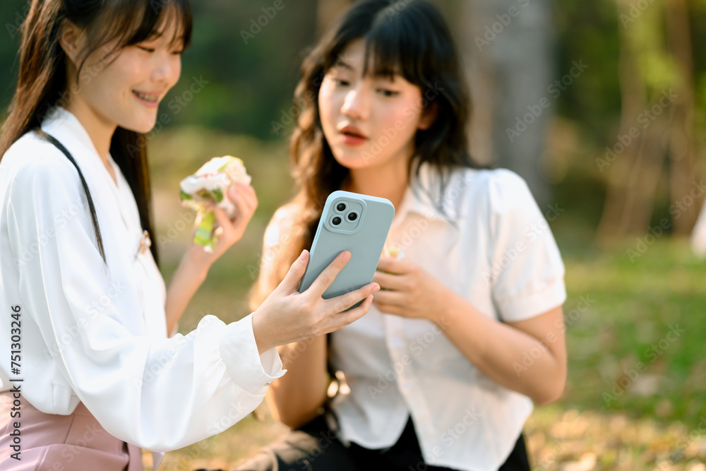 Shot of happy Asian female colleagues enjoying sandwich during lunch break and using mobile phone Shot of happy Asian female colleagues enjoying sandwich during lunch break and using mobile phone
