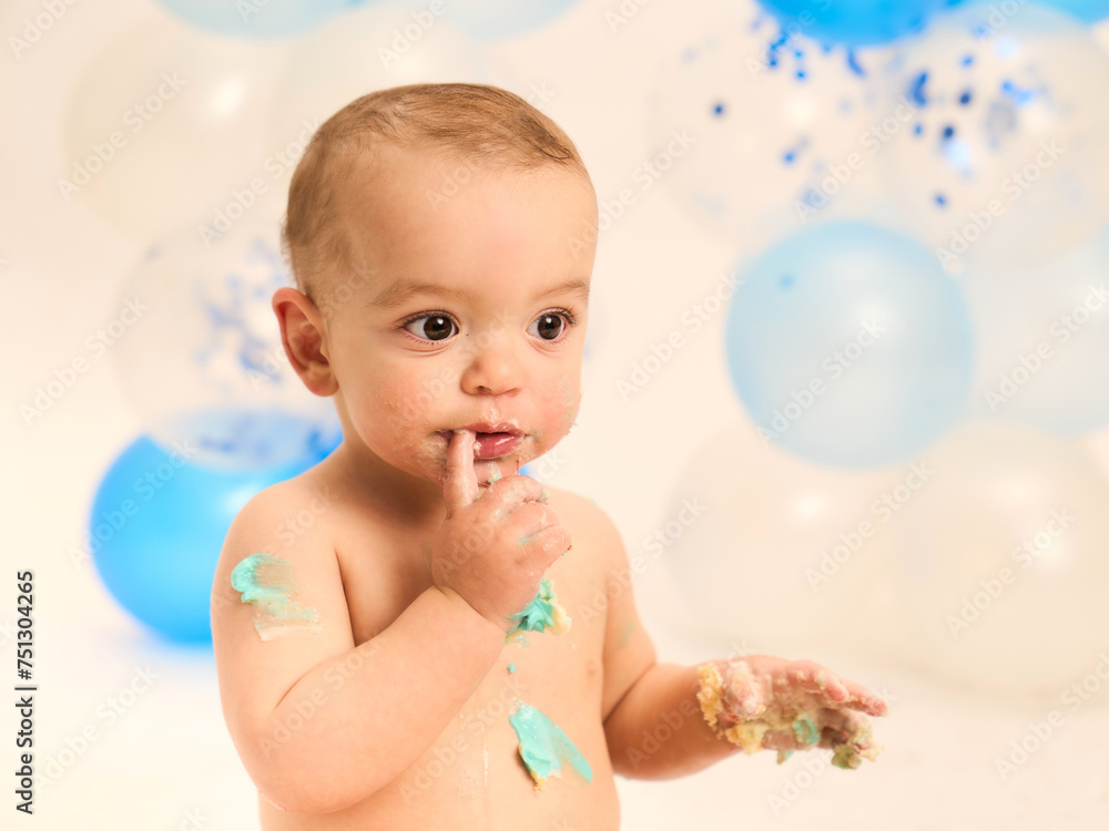 portrait of a beautiful baby with finger in mouth stained cake with balloons in background portrait of a beautiful baby with finger in mouth stained cake with balloons in background