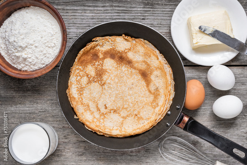 Frying pan with delicious crepes and ingredients for cooking on rustic wooden table. Thin pancakes in a pan with flour, eggs, milk and butter. Top view. Delicious breakfast