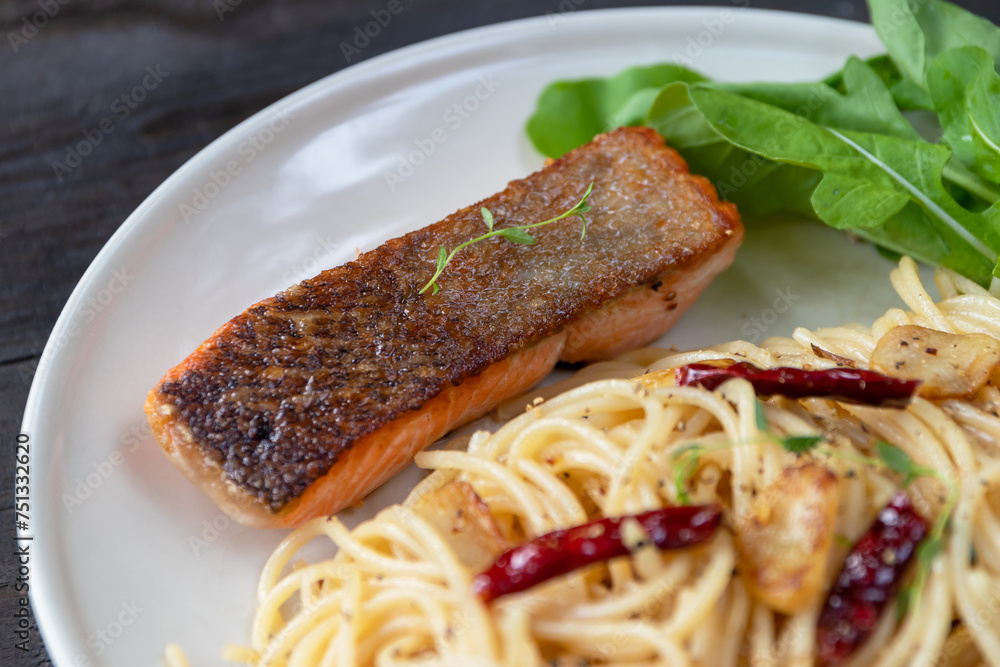 Spaghetti olio with salmon steak and herbs in white plate on dark wooden table background.