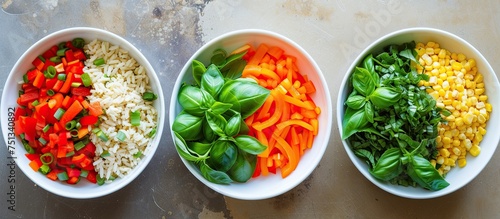 Three bowls on a table are filled with a variety of colorful vegetables, including basil, red bell pepper, carrot, corn, and brown rice. Each bowl holds a unique combination of fresh and vibrant