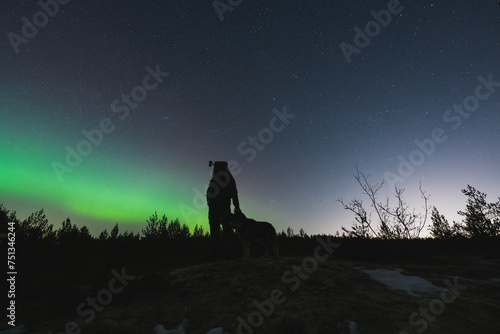 Silhouette of a man with a night vision device on his head and with a dog in nature at night against the backdrop of the starry sky and northern lights.