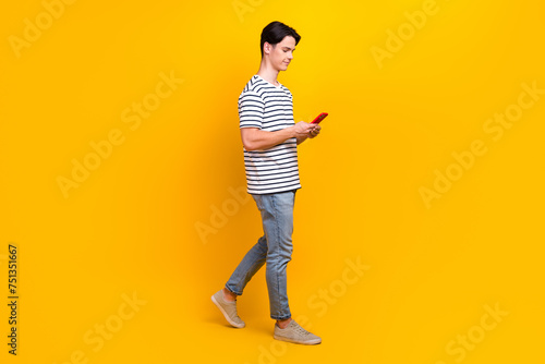 Full length photo of intelligent guy wear t-shirt look at smartphone read email walk to empty space isolated on yellow color background