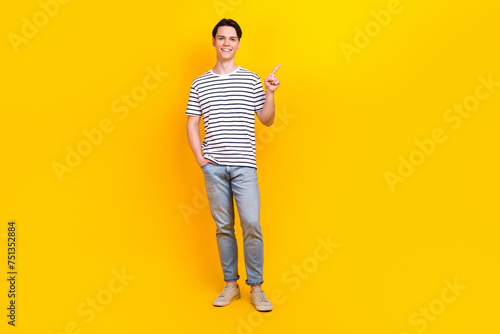 Full length photo of cheerful guy wear stylish t-shirt denim pants indicating at offer empty space isolated on yellow color background