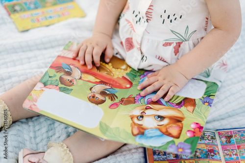 Portrait of beautiful daughter kid reading a book in summer nature. Closeup. Top view. Child playing toys in field at sunset. Cute baby girl sitting on a blanket in the park. Concept of family picnic.