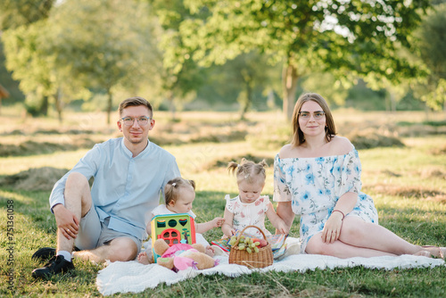 Happy young family in park on vacation together. Portrait of mom, dad, two daughter. Concept of family holiday. Father, mother, baby girls sitting on blanket on green grass, picnic in nature. photo