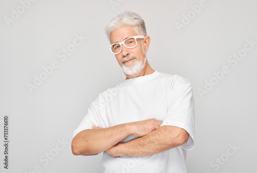 Confident senior man with glasses wearing white shirt arms crossed, standing on gray background.