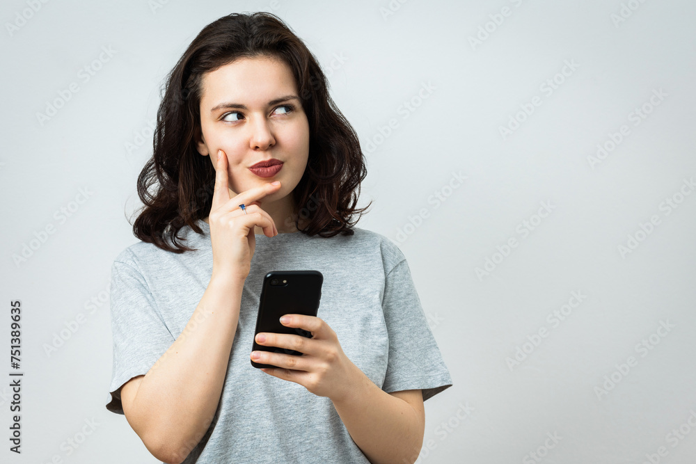 Pensive and smiling young caucasian woman attracted by some text or a product aside, holds mobile phone. Studio shot, isolated on light gray background Pensive and smiling young caucasian woman attracted by some text or a product aside, holds mobile phone. Studio shot, isolated on light gray background