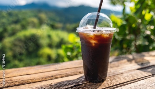closeup of take away plastic cup of iced black coffee americano on wooden table with green nature background
