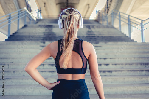 Photo of runner athlete standing in front of the stairs. Rear view shot of young woman standing by steps outdoors, holding bottle of water in hands down while listening to music.