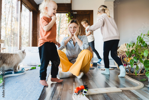 Fotografie Stressed out mother sitting in middle of toys while children naughty running around her at room