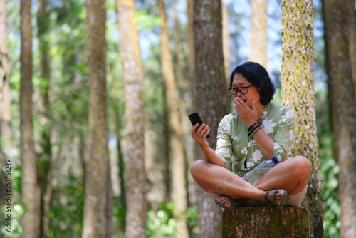 A woman sitting on a tree trunk is holding a smartphone while covering her mouth; crying, sad expression photo