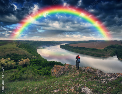 Tourist standing on the edge of the cliff and looking at the rainbow. Rainbow over the river. Nature of Ukraine © sergnester