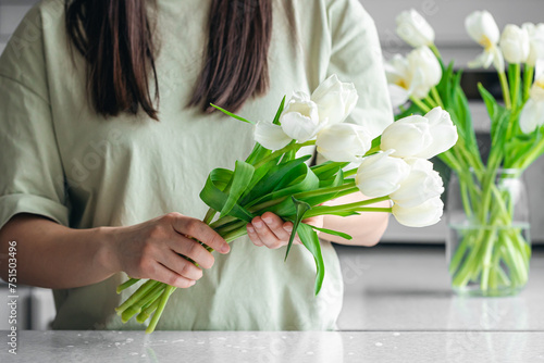 Woman putting bouquet of white tulip flowers into vase in the kitchen. #751503496