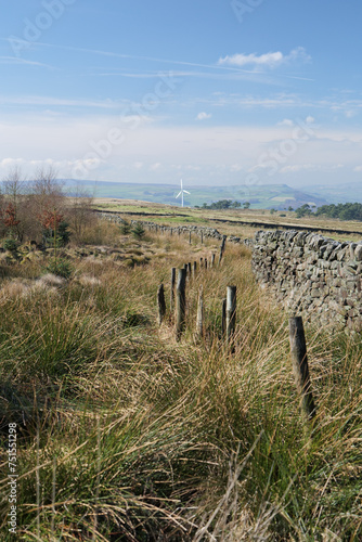 View of a Yorkshire landscape, with drystone wall, fence posts, and grasses. A wind turbine can be seen in the distance.