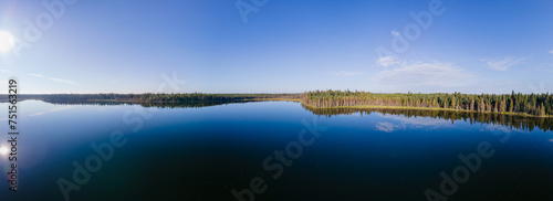 Panoramic aerial view of a large calm northern lake reflecting a clear blue sky.  The lakeshore is heavily treed with spruce and the sun reflects in the water on the left side of the image.
 photo