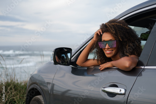 Portrait of a young black woman wearing sunglasses, leaning out of her car window during a relaxing getaway to the coast.