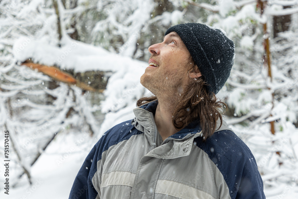 A man wearing a black hat and a blue and gray jacket is standing in the snow. He is smiling and looking up at the sky A man wearing a black hat and a blue and gray jacket is standing in the snow. He is smiling and looking up at the sky