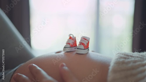 Pregnent woman hands caress her belly while a pair of small kid shoe is placed nearby. Mother holding baby in pregnant belly at home. Maternity prenatal care and woman pregnancy concept. photo