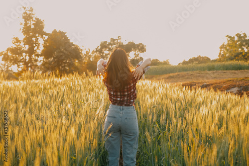 Young pretty woman in red summer dress and straw hat walking on yellow farm field with ripe golden wheat enjoying warm evening.