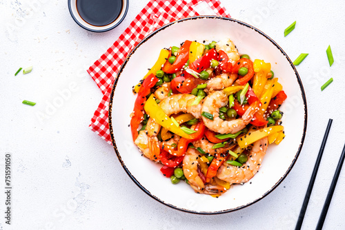 Spicy stir fry shrimps with colorful paprika, green peas, onion and sesame seeds with ginger, garlic and soy sauce in white bowl on kitchen table background, top view