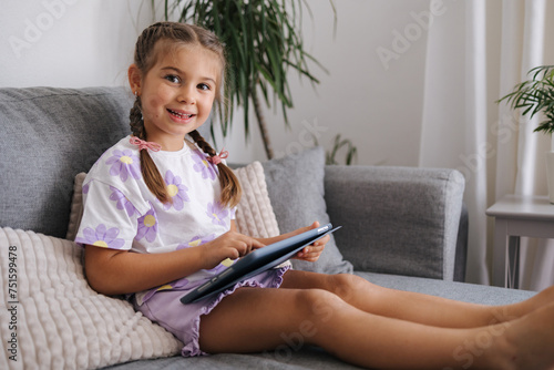 Cute little girl hold tablet in hands and look into camera. Girl sitting on sofa at home
