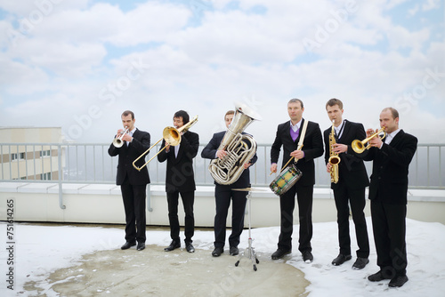 Brass band of six musicians in suits play on roof of tall building at winter