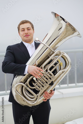 Man in black suit poses with tuba on roof of tall building at winter day